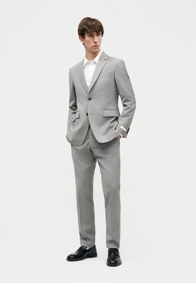 Νέος άνδρας που στέκεται με τα χέρια στις τσέπες, φορώντας ένα ανοιχτό γκρι κοστούμι, λευκό πουκάμισο και μαύρα παπούτσια επίσημα, μπροστά σε απλό φόντο.