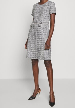 Frau trägt ein knieumspielendes, kurzärmliges schwarz-weiß gemustertes Kleid mit einem schmalen Gürtel, kombiniert mit schwarzen spitzen High Heels.