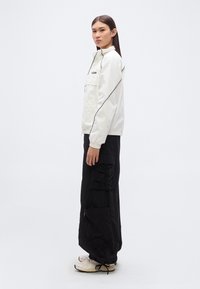 Λευκό ελαφρύ μπουφάν με μαύρες λεπτομέρειες στις ραφές, συνδυασμένο με μαύρο παντελόνι cargo με τσέπες και ρεβέρ. Το μοντέλο φοράει ανοιχτόχρωμα αθλητικά παπούτσια.