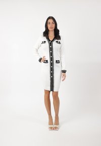 Abito a maglia bianco con dettagli neri, caratterizzato da scollatura a V, chiusura a bottoni sul davanti, due tasche e polsini a costine. Tessuto testurizzato con bordi a contrasto.