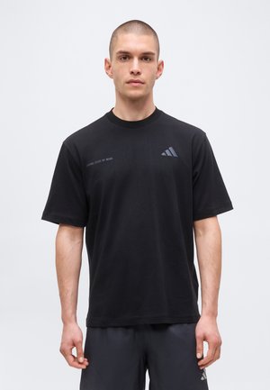 Muž s krátkými vlasy, který nosí černé tričko Adidas a šortky, stojí proti jednoduchému světle šedému pozadí a dívá se vpřed.