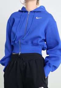 Sudadera con capucha corta azul con cierre de cremallera y cordones. Presenta un logo blanco de Nike en el pecho. Combinada con pantalones negros con cordones.