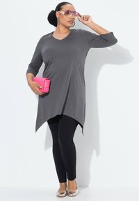 Harmaa pitkähihaista tunikaa, jossa on V-pääntie ja erikoismuotoinen helma, yhdistettynä mustiin legginsseihin ja kultaisiin ballerinatossuihin. Pitelee vaaleanpunaista laukkua.