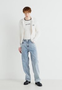 Chaqueta de punto blanco con nervios, camiseta negra con logo, vaqueros rectos de color azul claro y zapatos negros tipo slip-on, de pie contra un fondo en blanco.
