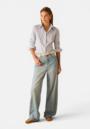 Donna che indossa una camicia a righe azzurro chiaro con bottoni, jeans slavati a gamba larga, cintura beige, mocassini marroni e gioielli in oro, in piedi con la mano in tasca.