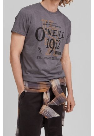 Mężczyzna ubrany w szarą koszulkę O'Neill 1952 z grafiką oraz brązowo-beżową koszulę w kratę zawiązaną wokół talii.
