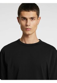 Melna T-krekls ar apaļu kakla izgriezumu un īsām piedurknēm. Audums izskatās gluds ar nedaudz brīvu piegriezumu. Nav redzamu rakstu vai grafikas.