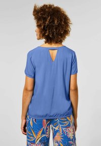 Blusa blu a maniche corte con dettaglio a chiave sul retro. Il tessuto è liscio e leggero, abbinata a pantaloni con fantasia.