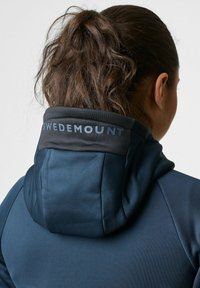 Hooded jack in donkerblauwe stof. Bevat een zwarte kraag met de tekst "SWEDEMOUNT". Glad materiaal, getailleerd ontwerp en zichtbare gestikte accenten.