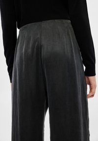 Calças de couro pretas com textura suave, apresentando um caimento relaxado e pregas na parte de trás, combinadas com um top preto de mangas compridas.