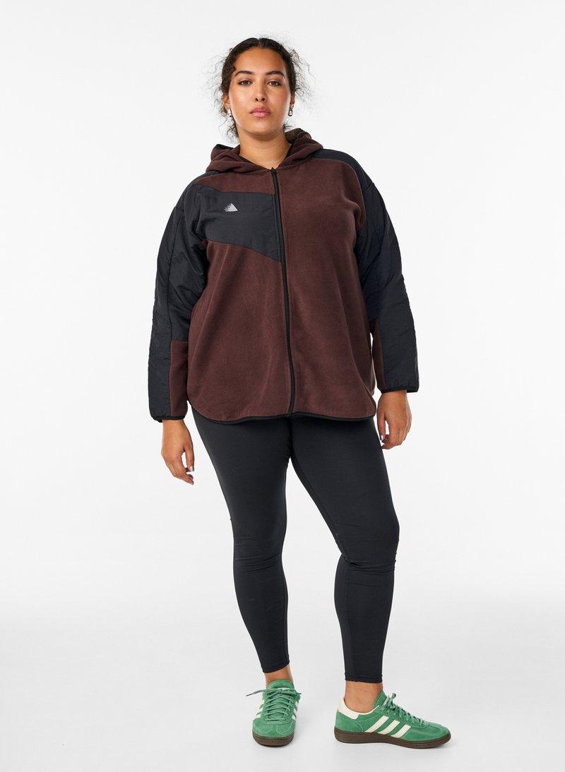Brown fleece zip-up jacket with black sleeves, hood, and logo. Styled with black leggings and green sneakers. Model poses facing forward.