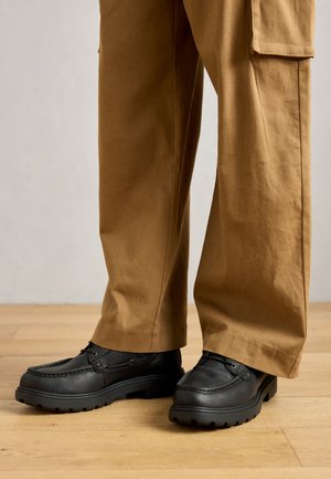 Black leather shoes with a chunky rubber sole, detailed stitching, and lace accents. Paired with tan, wide-legged trousers on a wooden floor.