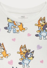 Witte geribde top van stof met kleurrijke cartoonhondjes in verschillende houdingen; lichte paarse en roze hartpatronen door het geheel. M&S-label zichtbaar.