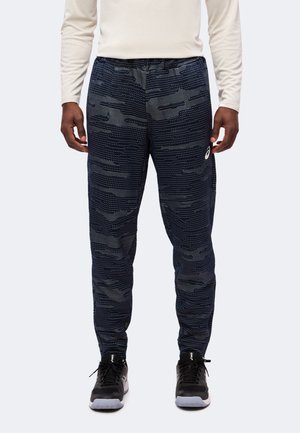 Modèle masculin portant un pantalon de sport noir et gris à motif avec une ceinture élastique et des chaussures de sport noires sur un fond uni.