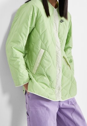 Person trägt eine hellgrüne gesteppte Nike-Jacke mit Druckknöpfen und hellviolette Cargohosen, die Hände in den Jackentaschen.