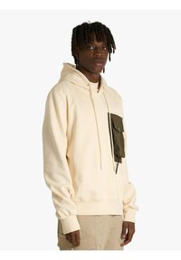 Felpa con cappuccio beige in cotone con cappuccio regolabile con coulisse. Presenta una tasca sul petto di colore verde scuro con chiusura a bottone. Polsini delle maniche con texture.