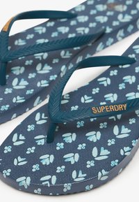 Tongs bleu marine avec motifs floraux bleu clair et brides texturées comportant la marque "SUPERDRY" en lettres orange.