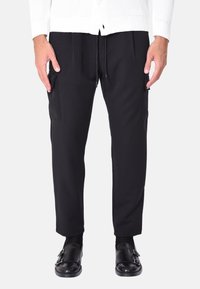 Pantaloni neri affusolati con vita regolabile, dotati di tasche laterali e una texture liscia. Abbinati a scarpe nere senza lacci.