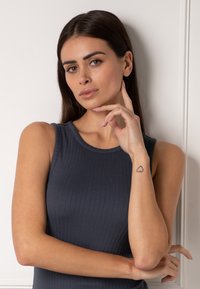Top senza maniche di un grigio scuro a coste con scollatura rotonda. Il modello posa con una mano vicino al viso, rivelando un piccolo tatuaggio a forma di cuore sul polso.