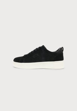 Sort ruskind lavtop sneaker med hvid tyk gummisål og sorte snørebånd på en ensfarvet lys baggrund.