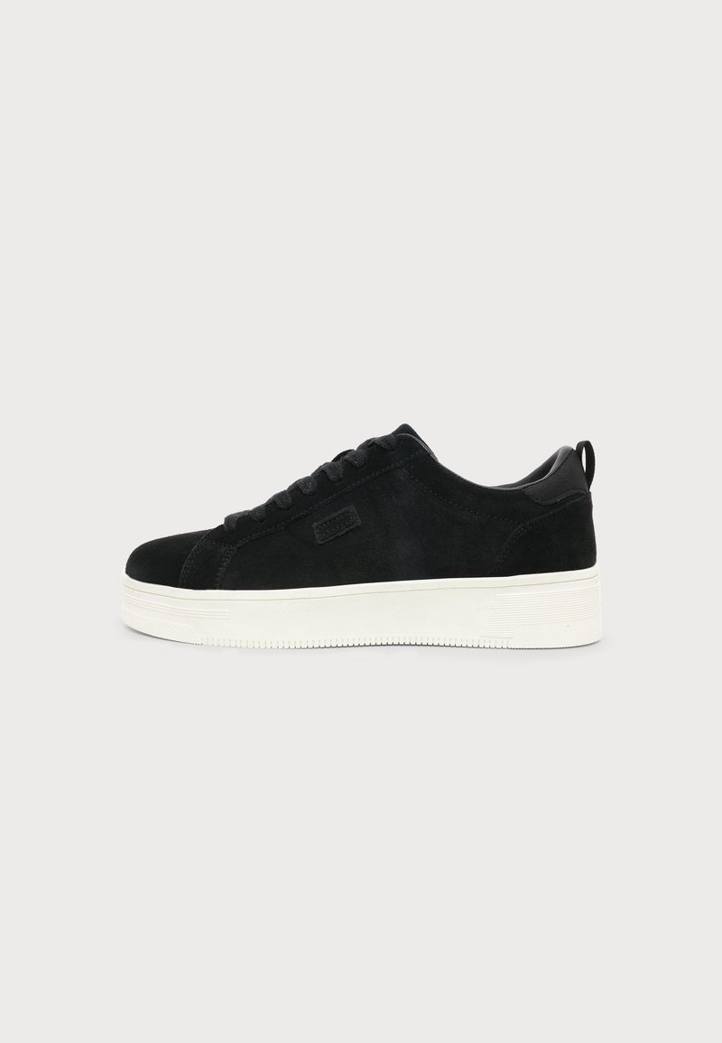 Μαύρο καστόρινο χαμηλό sneaker με λευκή παχιά λαστιχένια σόλα και μαύρα κορδόνια σε απλό ανοιχτό φόντο.
