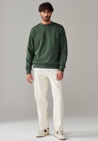 Grüner Crewneck-Pullover aus weichem Stoff, kombiniert mit weißen, geraden Hosen und lässigen Sneakers mit braunen Akzenten. Einfaches, minimalistisches Design.