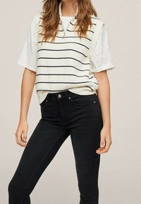 Chaleco de punto color crema con rayas horizontales azul marino, mangas cortas y cuello con cremallera, combinado con jeans skinny negros.