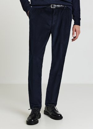 Pantaloni in velluto a coste blu navy con sottili righe verticali, pieghe frontali, tasche laterali e cintura in pelle nera. Indossati con scarpe nere stringate.