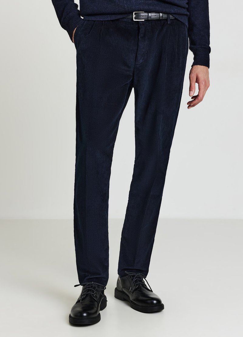 Marineblaue Cordhosen mit Nadelstreifen, vorderen Falten, seitlichen Taschen und einem schwarzen Ledergürtel. Kombiniert mit schwarzen Schnürschuhen.