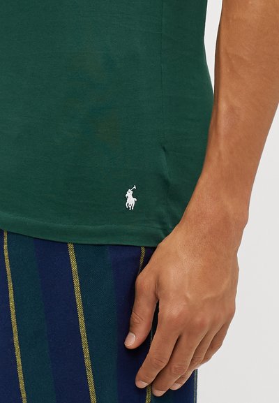 Žalias medvilninis marškinėliai su balta siuvinėjimu logotipu apačioje, vilkintis su mėlynai ir žaliai juostuotais šortais bei matoma ranka.