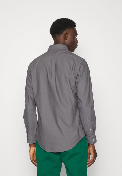 Camisa gris de manga larga con collar abotonado, corte ajustado y pliegues en la parte posterior. La tela parece lisa con un ligero brillo.