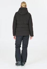 Doudoune noire avec capuche, présentant des sections matelassées et un design ajusté, assortie à un pantalon noir et des chaussures sombres.