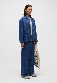 Jeansjacke und weit geschnittene Hose mit floralem Muster, beide in Blau. Graues Shirt darunter, hält eine beigefarbene Tragetasche mit schwarzem Text.