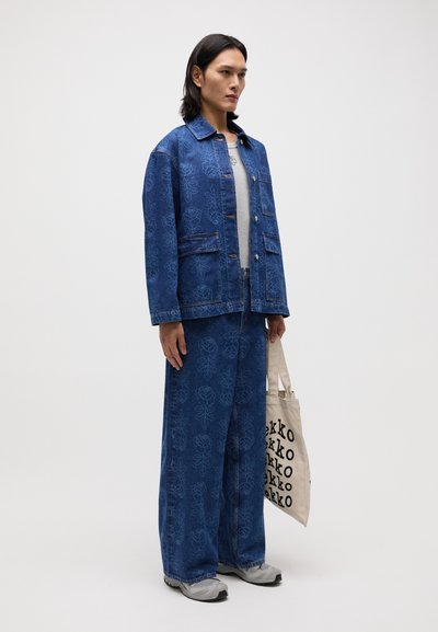 Veste en jean et pantalon large avec un motif floral, tous deux en bleu. Chemise grise en dessous, tenant un sac fourre-tout beige avec du texte noir.