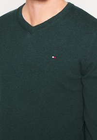 Homme portant un pull vert foncé à col en V avec un petit logo rectangulaire rouge, blanc et bleu sur la poitrine gauche.