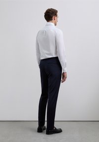 Camisa blanca de manga larga con abertura trasera, combinada con pantalones azul marino oscuro con bolsillos traseros y zapatos negros sin cordones.