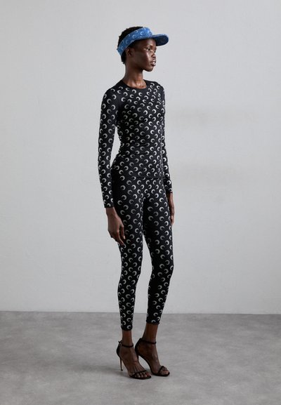 Black fitted bodysuit and leggings with a repetitive silver crescent pattern; paired with a blue fabric visor and black high-heel sandals.