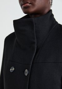 Černý kabát s vysokým límcem, dvěma velkými knoflíky a hladkou texturou. Má tvarované švy a moderní design.