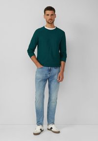 Teal langemouw shirt met textuur en een witte ronde hals, gecombineerd met lichtblauwe jeans en witte sneakers met zwarte accenten. Casual fit.