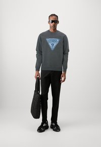 Sudadera gris oscuro con un logo triangular azul, pantalones negros, sosteniendo un bolso acolchado y usando gafas de sol negras y zapatillas deportivas.