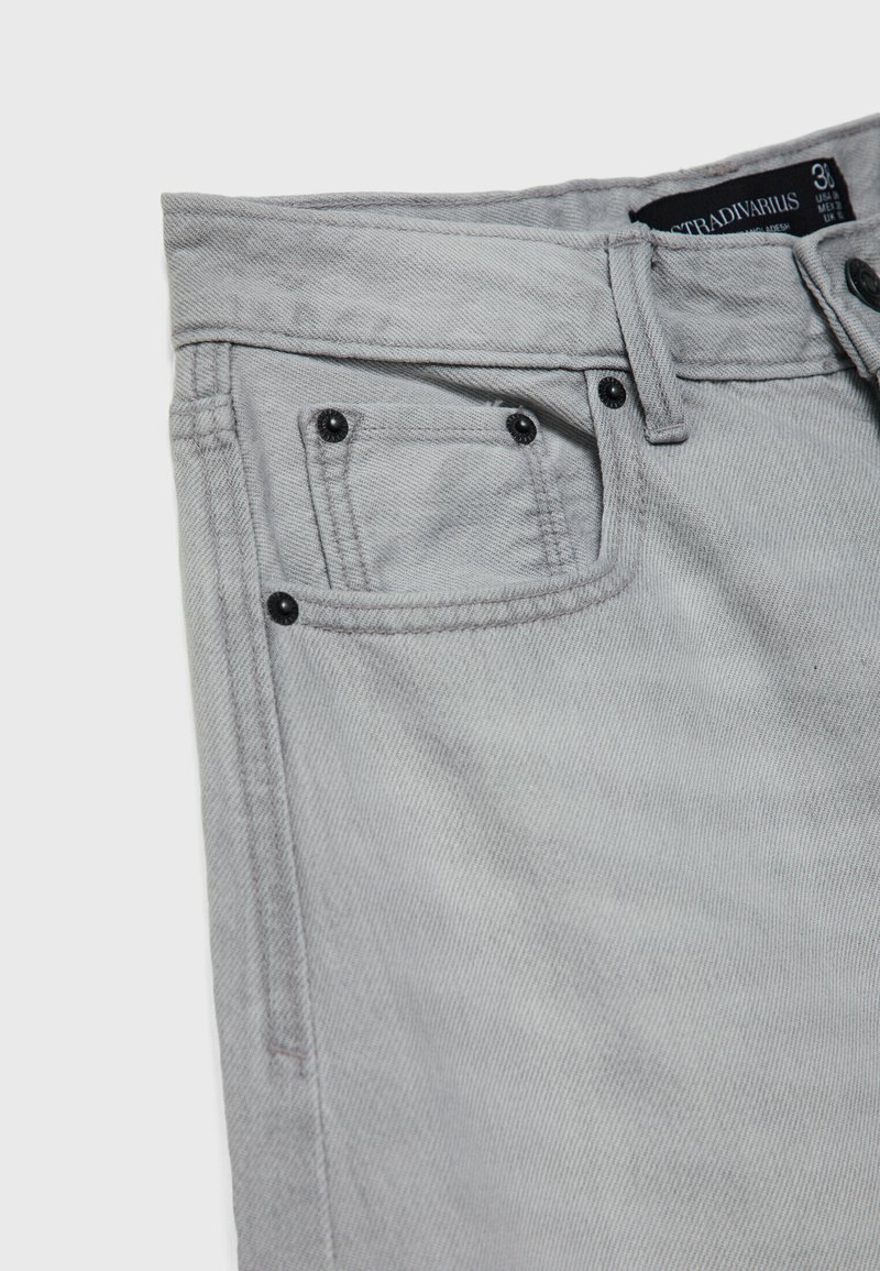 Section de jeans en denim gris clair montrant une poche avant avec une petite poche à pièces, des rivets noirs, une passante de ceinture et une étiquette de taille partiellement visible.
