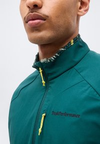 Κοντινό πλάνο ενός άνδρα που φοράει ένα πράσινο μπουφάν PeakPerformance με κίτρινη φερμουάρ και φθαρμένο γιακά.