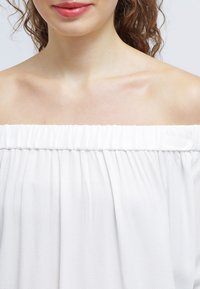 Top blanco sin hombros con escote fruncido, hecho de una tela ligera, que presenta una textura suave y un diseño fluido.