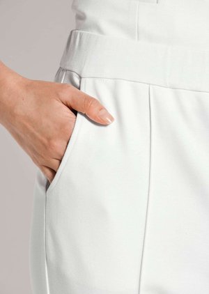 Main avec des ongles naturels placée dans la poche latérale d'un pantalon blanc taille haute avec une large ceinture.