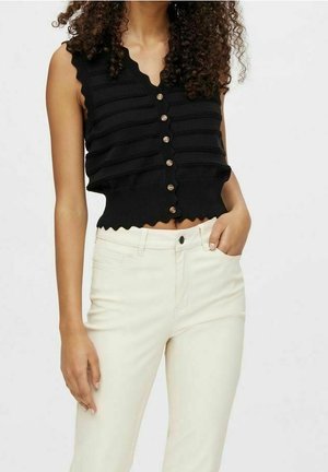Frau trägt ein ärmelloses schwarzes, gestricktes Top mit Knöpfen, gezacktem Saum und hoch taillierte, offwhite Hose, Hand in der Tasche.