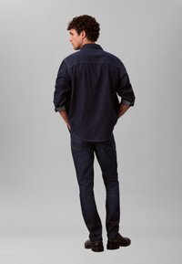 Τζιν πουκάμισο σε σκουρό μπλε χρώμα, με ριγμένα μανίκια και κυρτή γραμμή στο κάτω μέρος. Συνδυάζεται με σκουρό μπλε τζιν παντελόνια και μαύρα παπούτσια, σε όψη από πίσω.