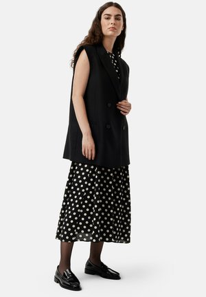 Black sleeveless blazer over a black and white polka dot dress, paired with shiny black loafers. Smooth textures and tailored fit.
