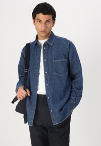 Camisa de mezclilla azul con botón en la parte delantera, un solo bolsillo en el pecho y mangas largas. Llevada sobre una camiseta blanca, combinada con pantalones oscuros.