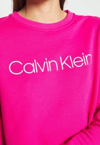 Felroze sweatshirt met een ronde hals en een witte "Calvin Klein"-logo gedrukt op de borst. Zachte stof met een gladde textuur.