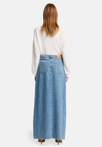 Gonna maxi in denim leggera di colore blu chiaro con vita alta, due tasche posteriori e un'etichetta in vita. Indossata con una maglietta bianca a maniche lunghe.